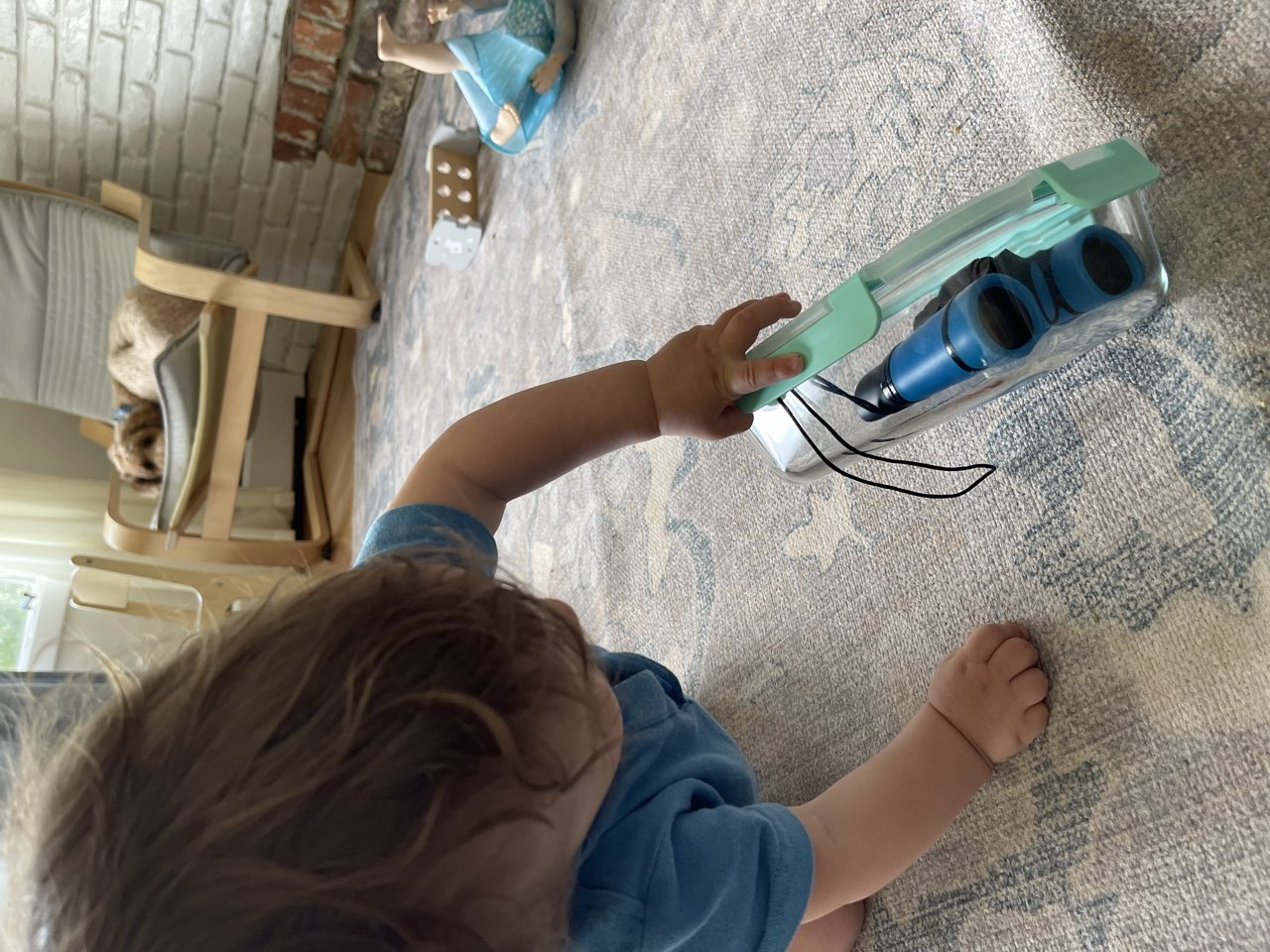 A toddler in a blue shirt reaches across a rug to grasp a toy with a teal handle.